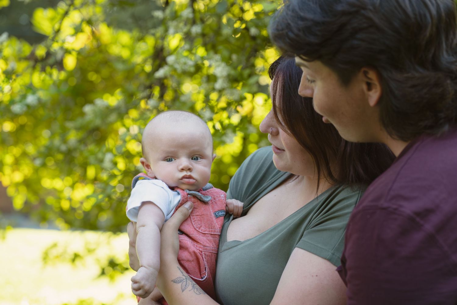 A small baby being held outdoors with sunlit green foliage in the background.