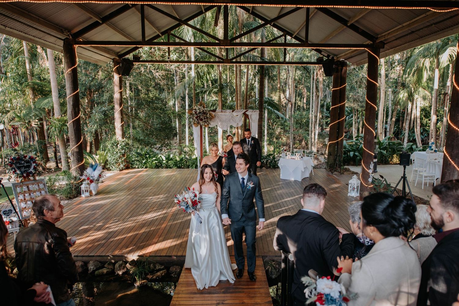 Newlyweds walking down aisle while guests celebrate in rustic outdoor venue.