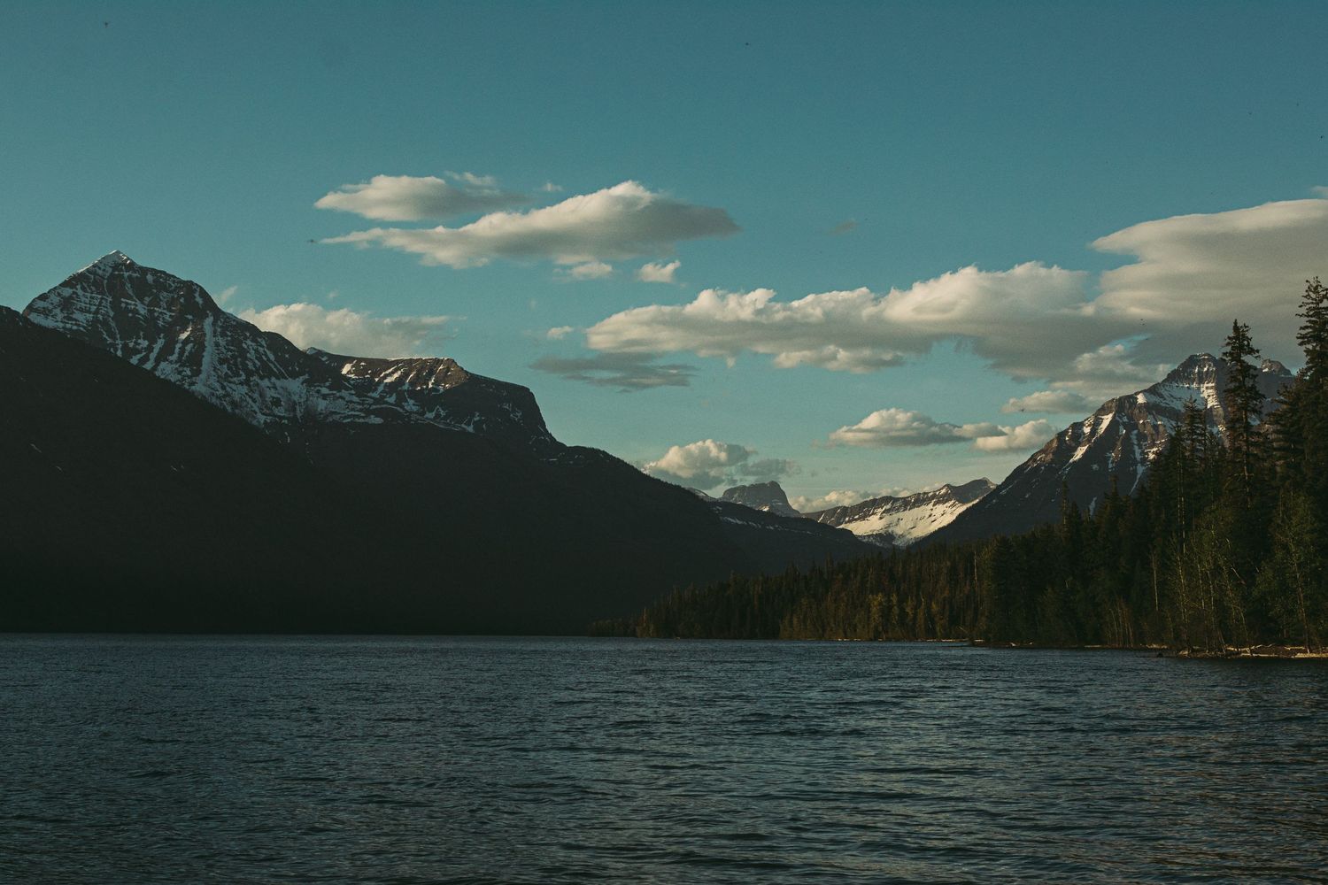 Scenic mountain lake landscape with snow-capped peaks and fluffy clouds in the distance.