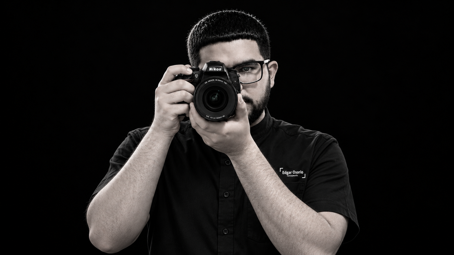 Professional photographer in black shirt holding DSLR camera against dark background in black and white.