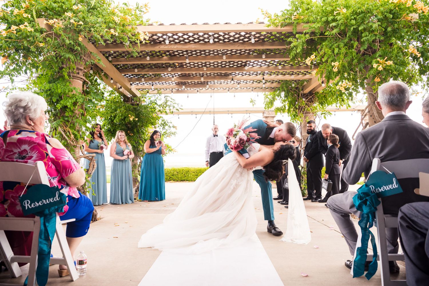A wedding ceremony under a vine-covered pergola with guests watching the couple.