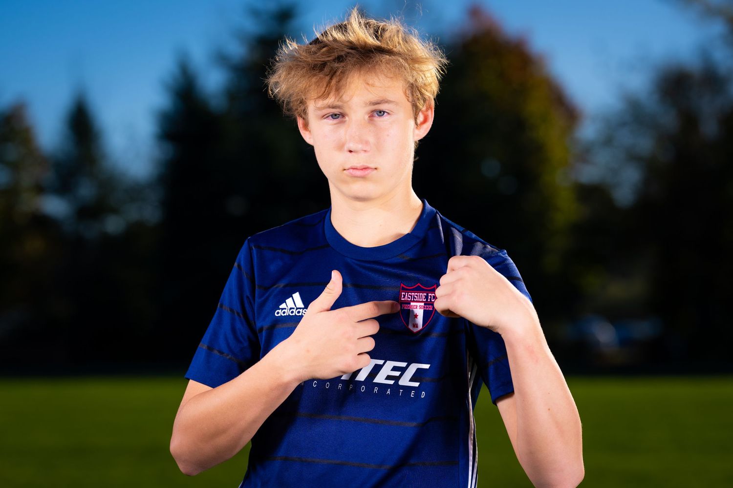 A male soccer player from Eastside FC Premier Soccer pointing at the team crest on his jersey.