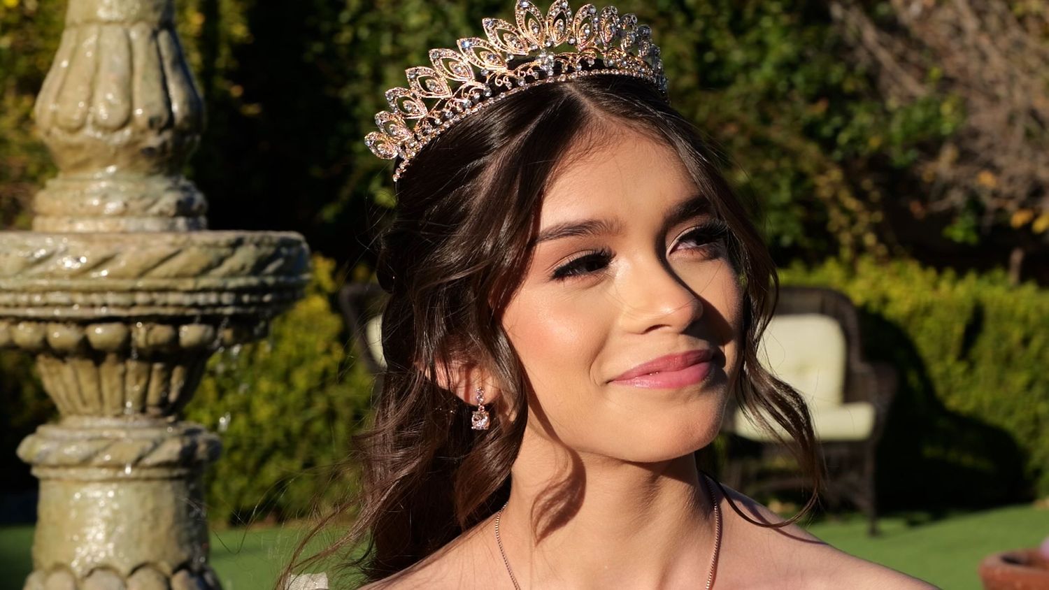 Warm sunlit portrait showcasing delicate crown and jewelry against stone backdrop.