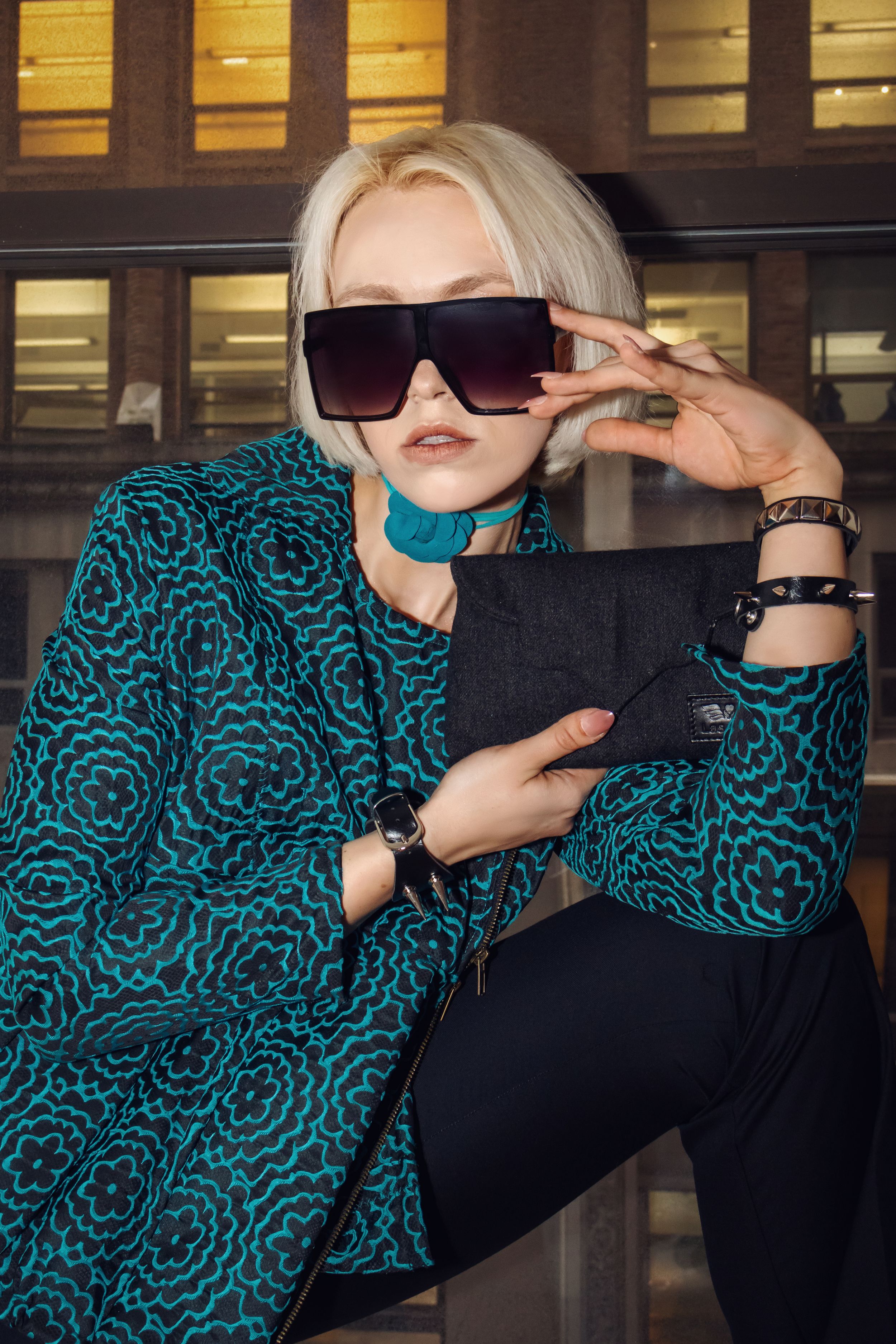 Stylish winter fashion featuring a turquoise patterned coat, black sunglasses, and dark clothing against an urban backdrop.