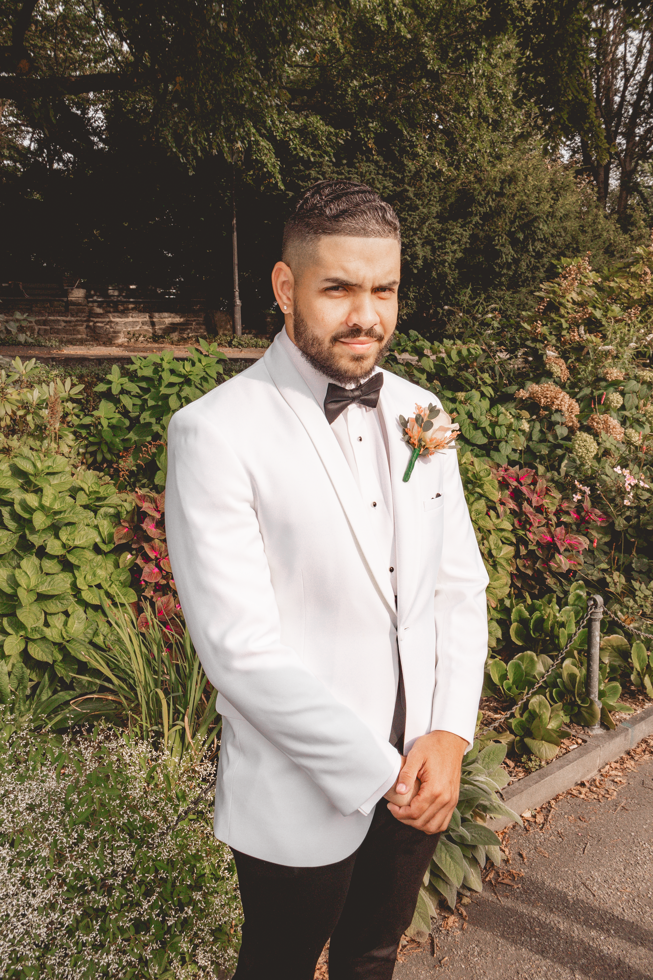 Groom in white tuxedo jacket and black bow tie poses in garden setting.