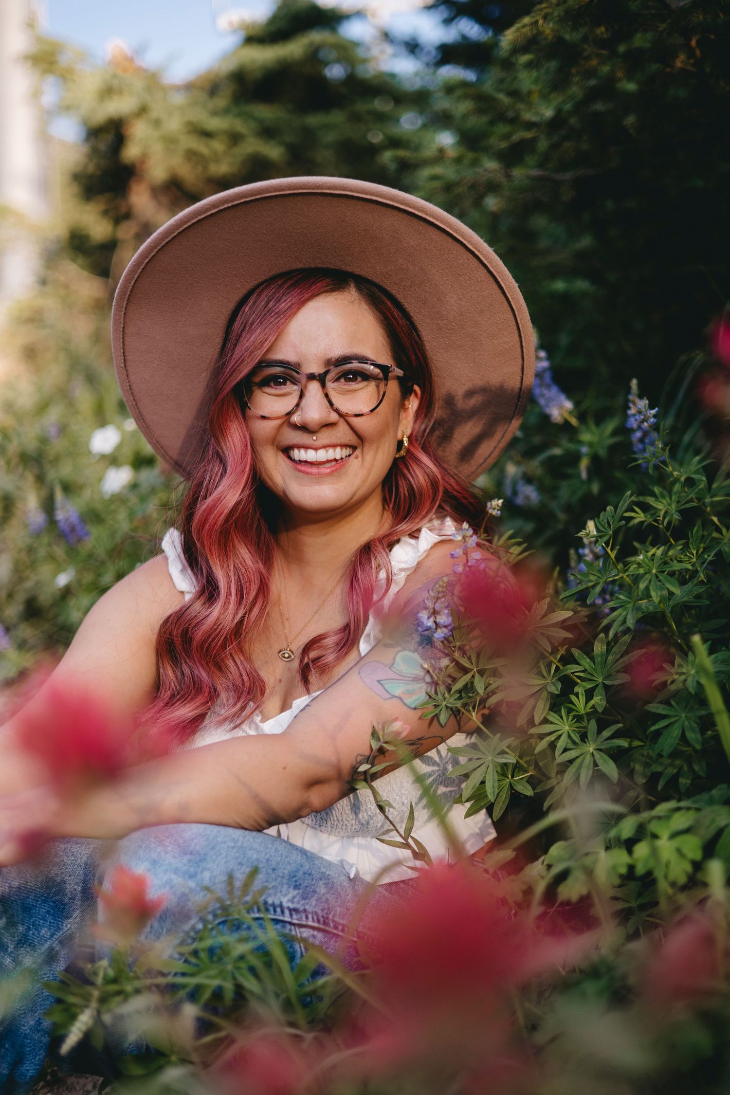 woman with pink hair, wearing glasses, a tan hat, gold hoop earrings, and an evil eye, opal necklace, sitting amongst wild flowers