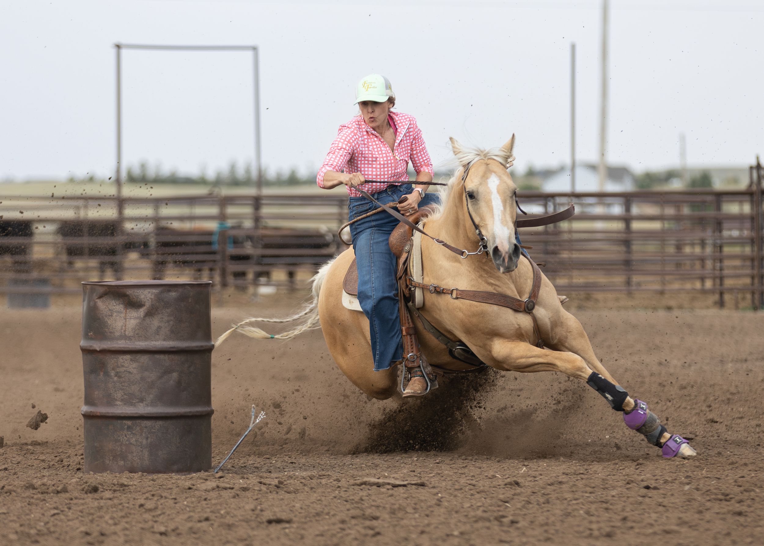 Woman in pink shirt rides palomino horse around barrel in dusty rodeo arena during barrel racing competition.