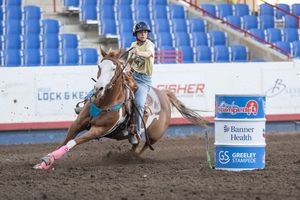 Young rider competes in barrel racing at Greeley Stampede rodeo, horse leaning into sharp turn around red barrel.