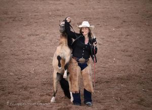 Young rider in western attire poses with a tan horse in a dirt arena.