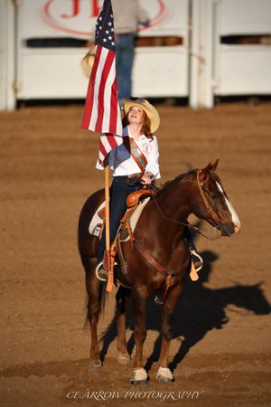 A rider carries the American flag while mounted on a brown horse in a rodeo arena.