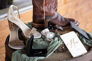 Wedding flat lay with bridal heels, cowboy boots, rings, and a gold calligraphy Her vows book on rustic wood.