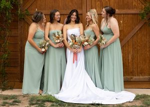 Bride in white gown with four bridesmaids in sage green dresses holding bouquets, posing in front of a wooden barn door.