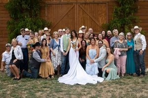Large wedding party posing together outdoors in front of rustic barn doors, featuring bride in white gown and guests in western attire.
