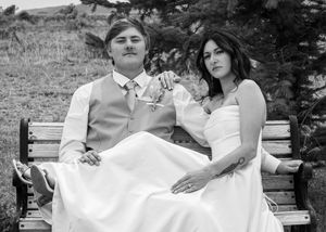 Black and white photo of a bride and groom sitting on a wooden bench outdoors on their wedding day.