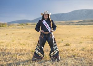 Woman in cowboy hat and Arkansas sash wearing decorative chaps poses in golden field with mountain backdrop.