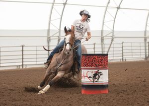 Barrel racer on a gray horse making a tight turn around a decorated barrel in an indoor rodeo arena.