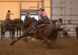 Barrel racing competition at an indoor arena with a horse and rider making a sharp turn in dusty conditions.