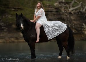 Woman in lace dress riding bareback on black horse in water.