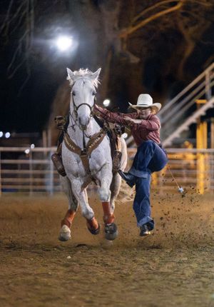A cowboy performs a dismount from a galloping white horse during a nighttime rodeo competition.