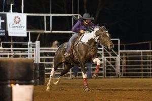 Rodeo rider competes in bronc riding event at night arena with illuminated dirt ground and metal fencing.