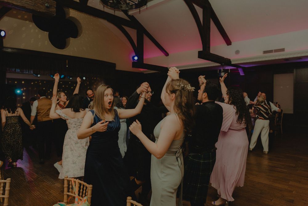 Guests dancing joyfully at a wedding reception in a decorated venue with colorful lights.
