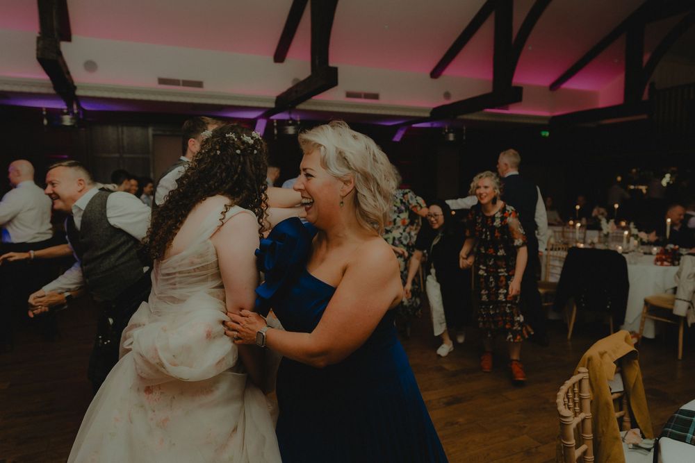 Bride in white gown shares a joyful embrace with a guest in a blue dress on the wedding reception dance floor.