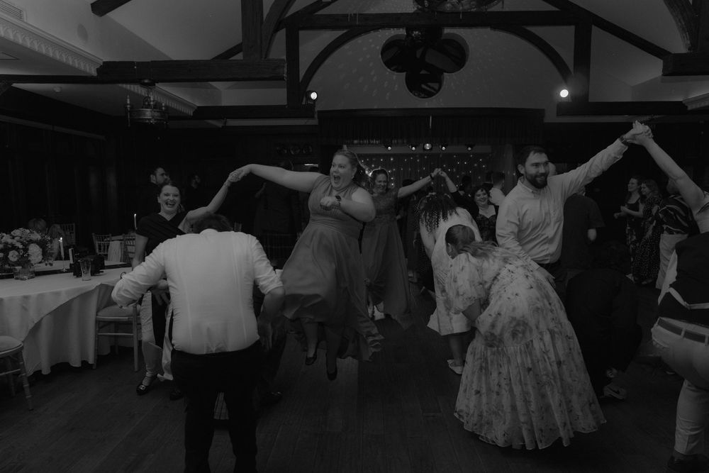 Black and white photo of wedding guests dancing together in a circle at a reception venue.