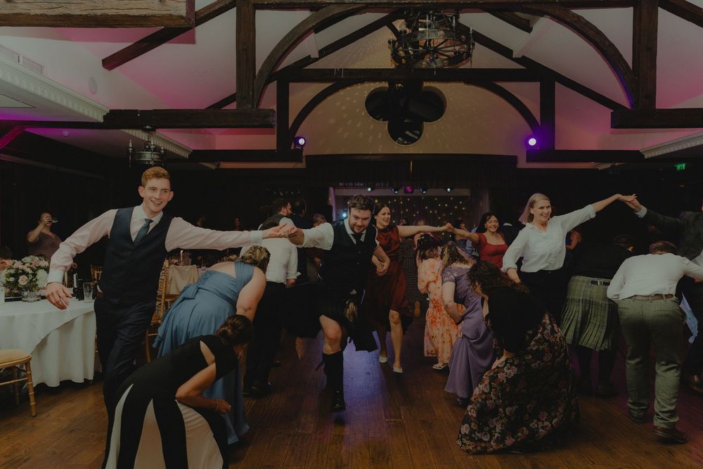 Wedding guests energetically dancing in a rustic venue with wooden beamed ceiling and purple lighting.