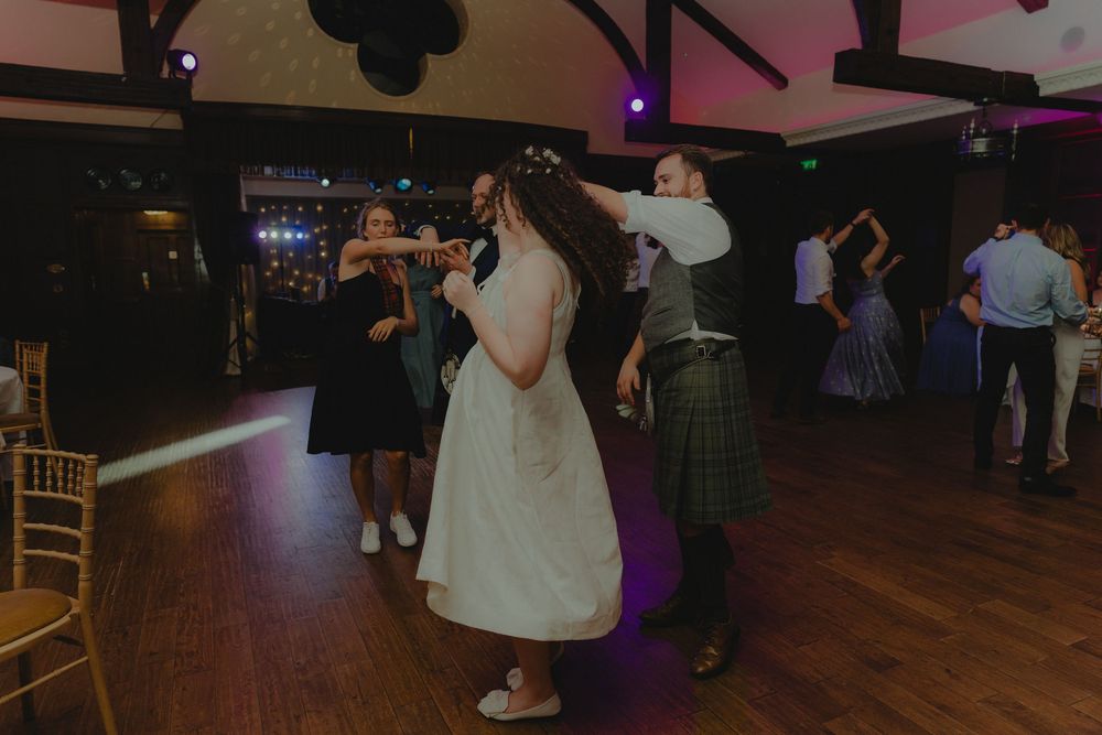 Bride in white dress dances with guests in kilts on a wooden dance floor at a Scottish wedding reception.