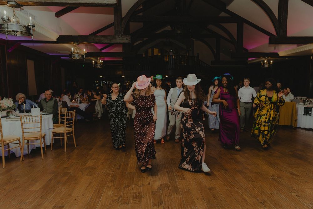Wedding guests in colorful gowns and hats line dancing at a reception in a rustic ballroom.