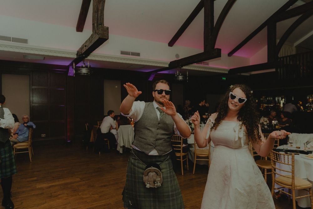 Bride and groom dancing joyfully in sunglasses at Scottish wedding reception, groom wearing traditional tartan kilt.