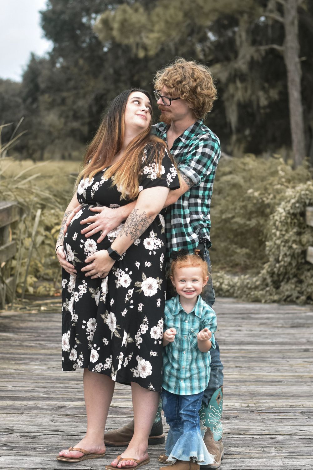 Expecting family poses together on wooden boardwalk surrounded by nature.