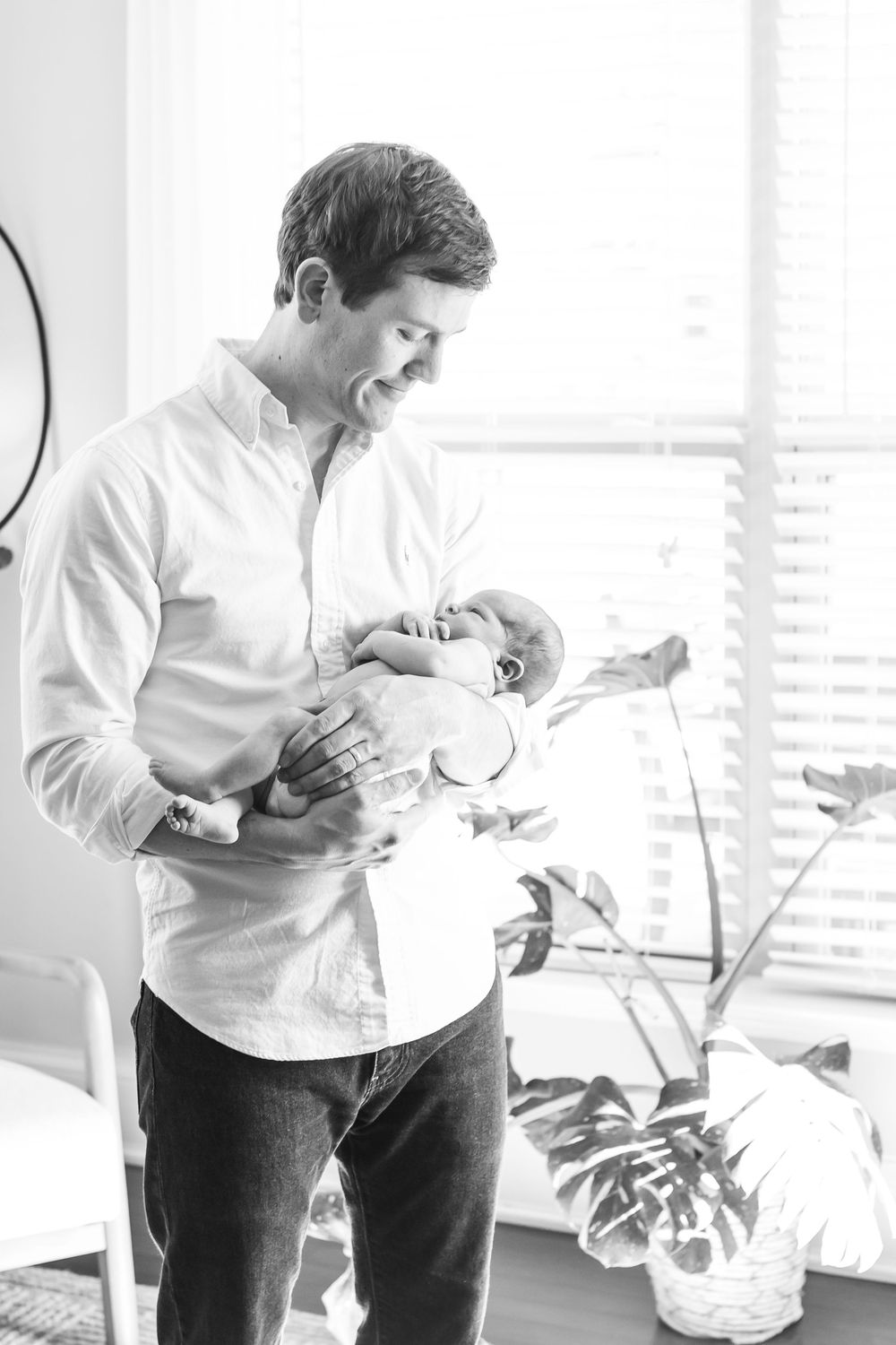 A black and white photo of a smiling father standing by a bright window, gently cradling his newborn baby.