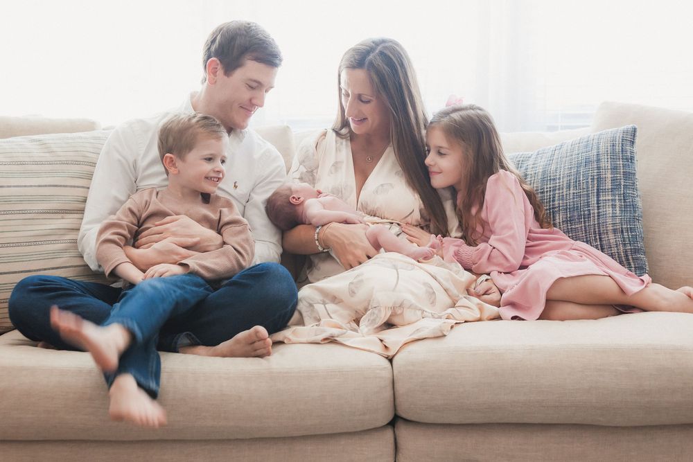 A happy family of five sits together on a beige sofa, parents and two children admiring their newborn baby.