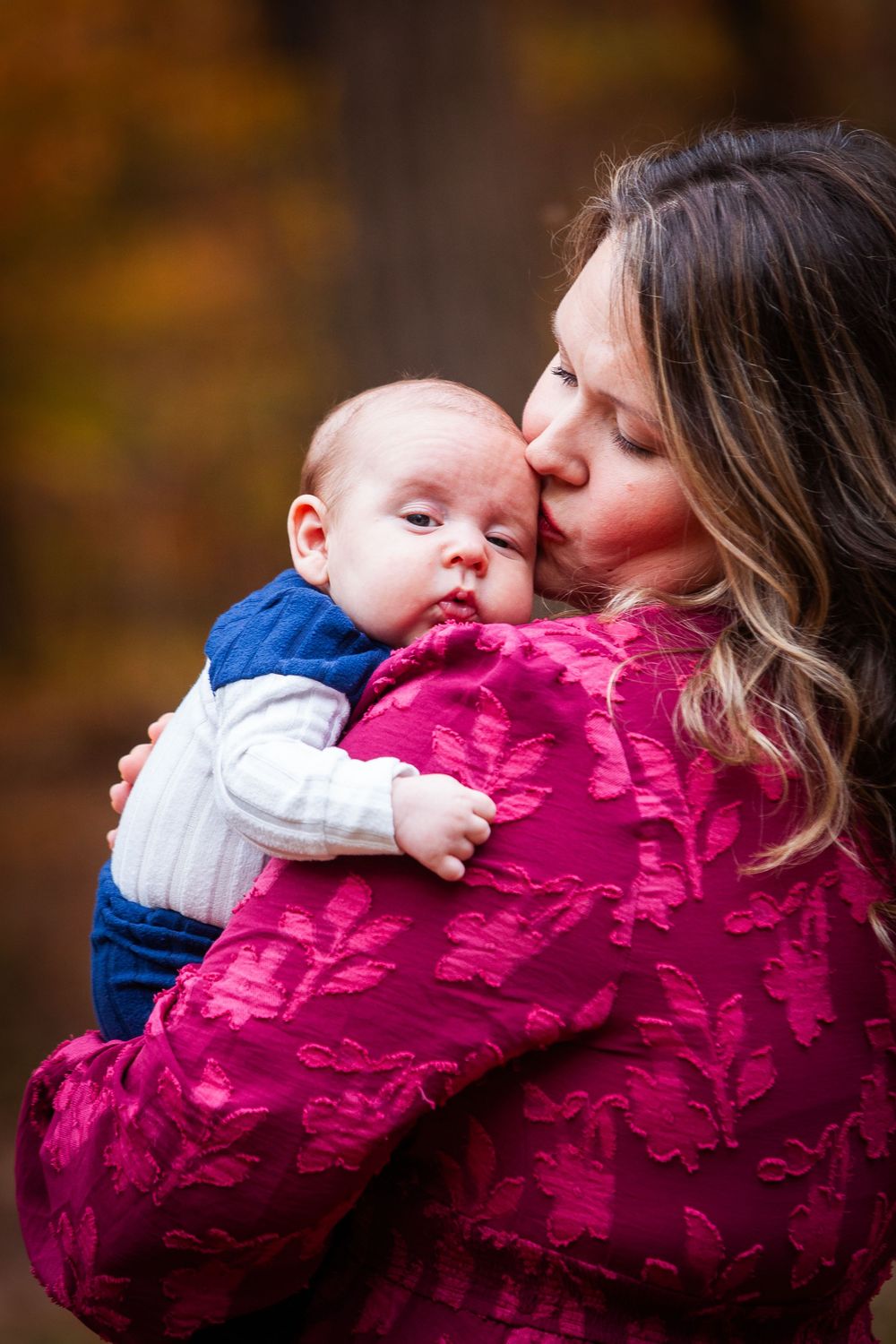 Mother in pink jacket tenderly kisses her infant baby outdoors in autumn setting.