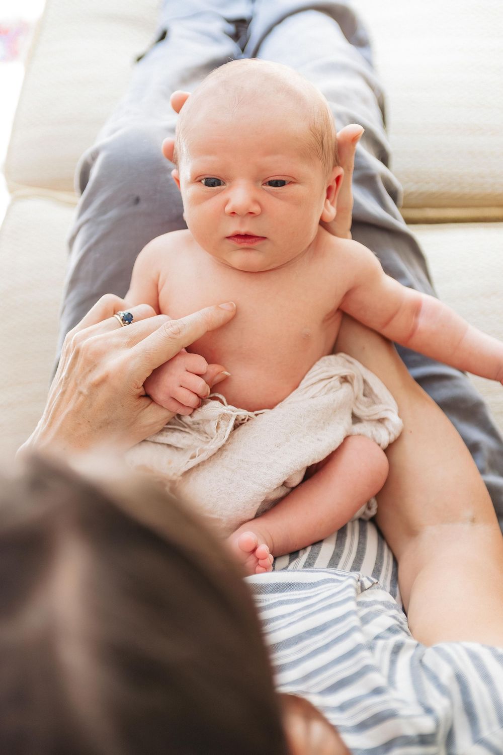 A newborn baby lies alert and undressed on a parent's lap, being gently supported by caring hands.