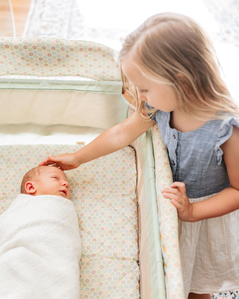 Young girl gently touches a swaddled newborn baby lying in a pastel patterned bassinet.