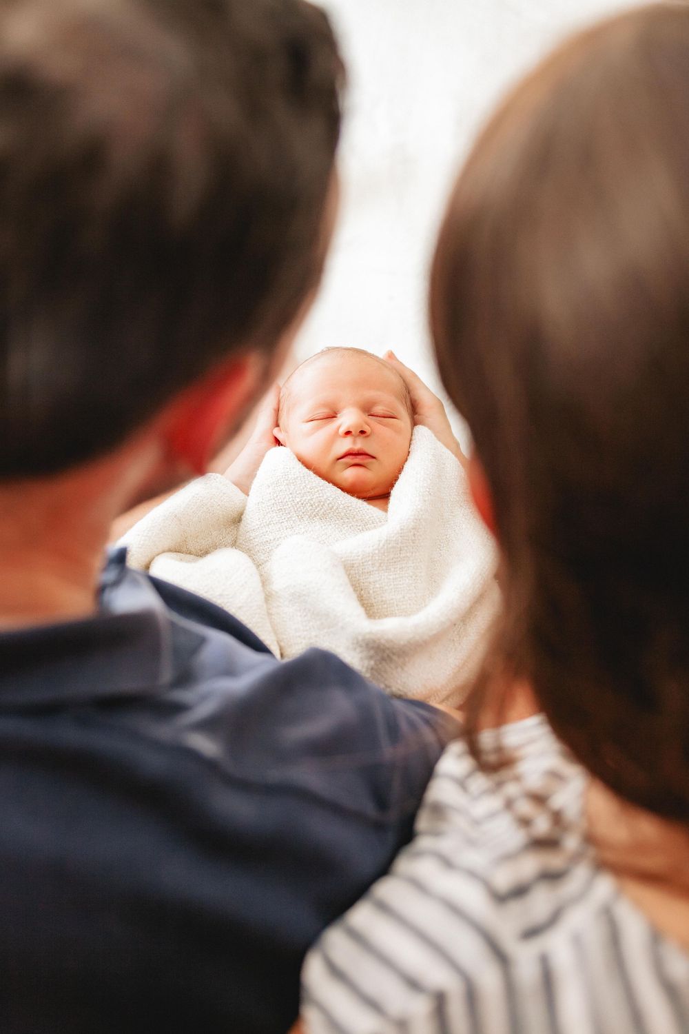 Two parents lovingly gaze at their sleeping newborn baby wrapped in a white blanket.