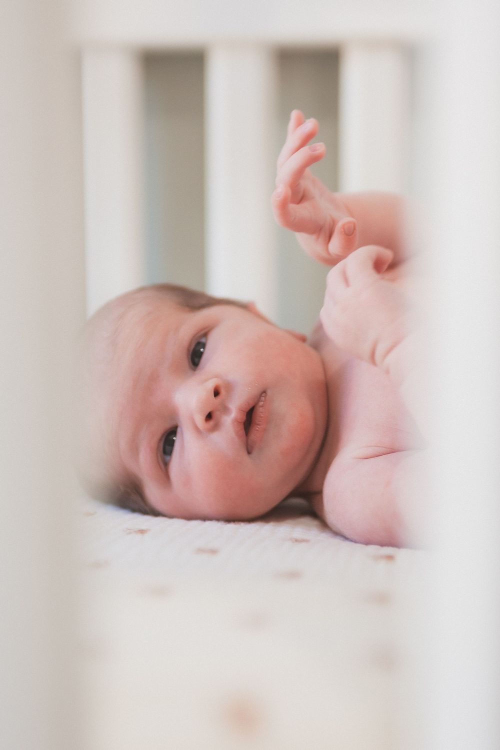 An alert newborn baby lies in a white crib, reaching one tiny hand upward in a softly lit nursery.