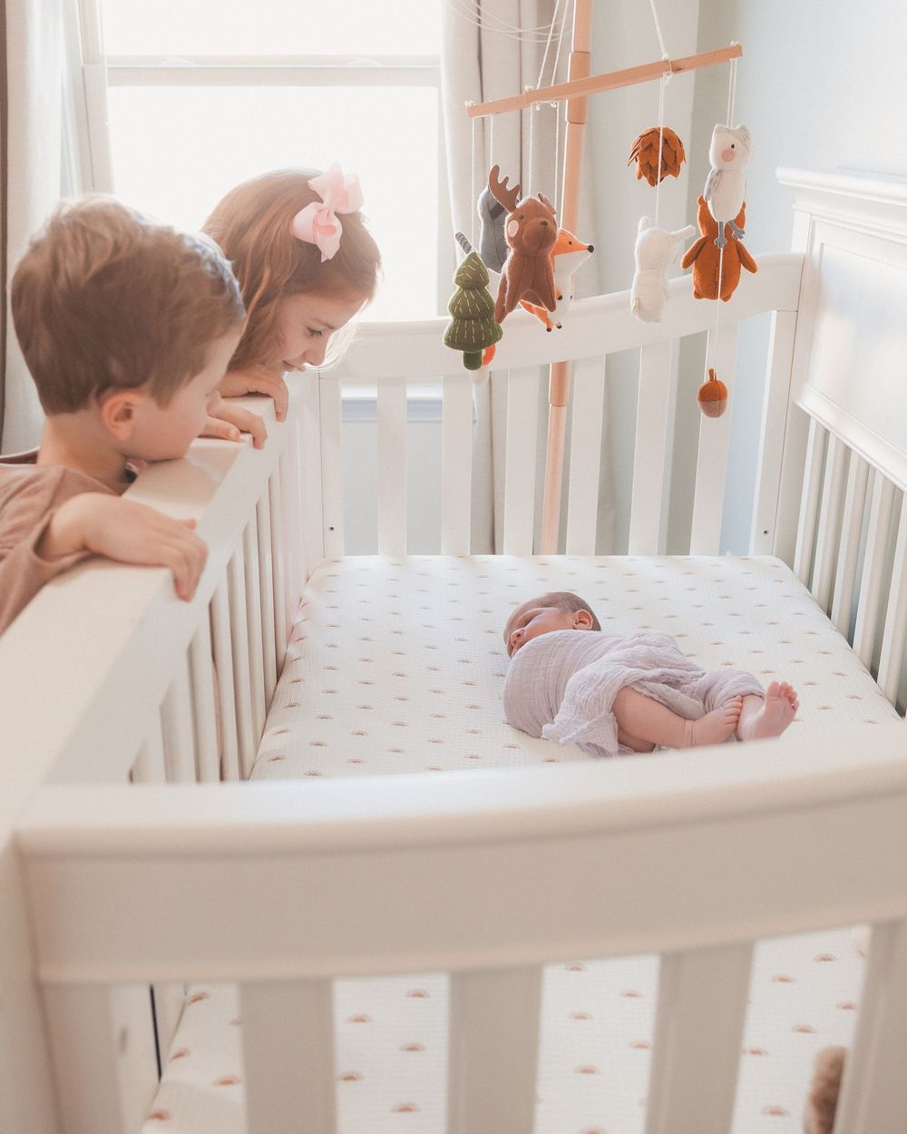 Two young siblings lean over a white crib to look at their sleeping newborn baby with a woodland animal mobile above.