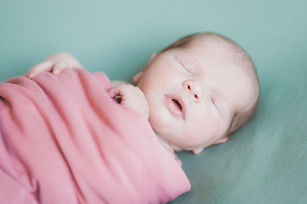 A peaceful newborn baby sleeps swaddled in a soft pink blanket on a mint green background.