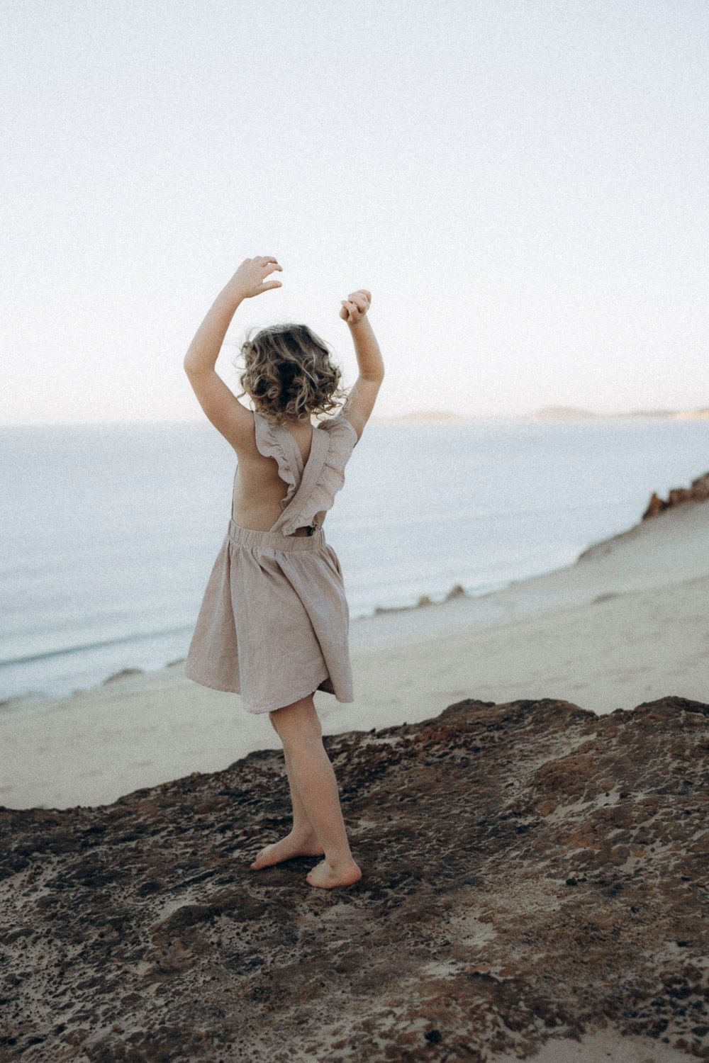 A person in a beige dress dances freely on a coastal cliff overlooking the ocean at dusk.