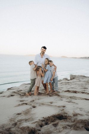 A family stands together on rocky coastal terrain at sunset while wearing coordinated light-colored casual outfits.