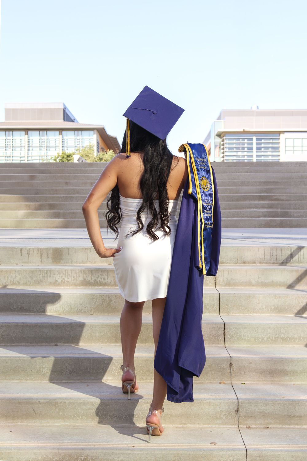 Graduate in cap and white dress holding gown, viewed from behind on steps.