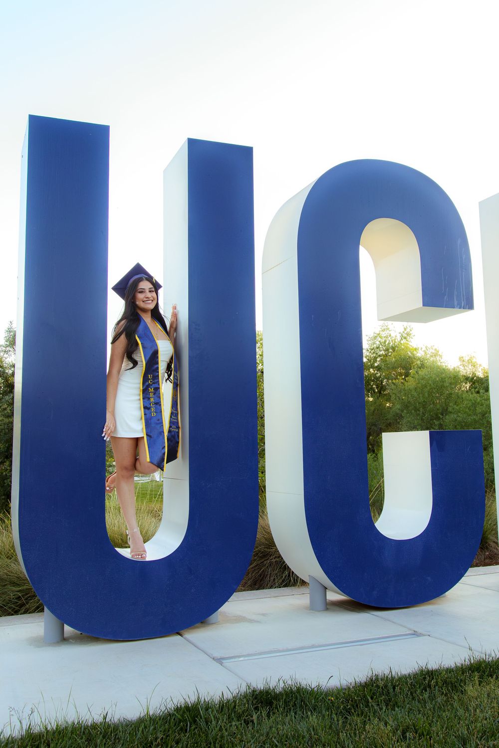 Smiling graduate posing between large UC letter sculptures on campus.