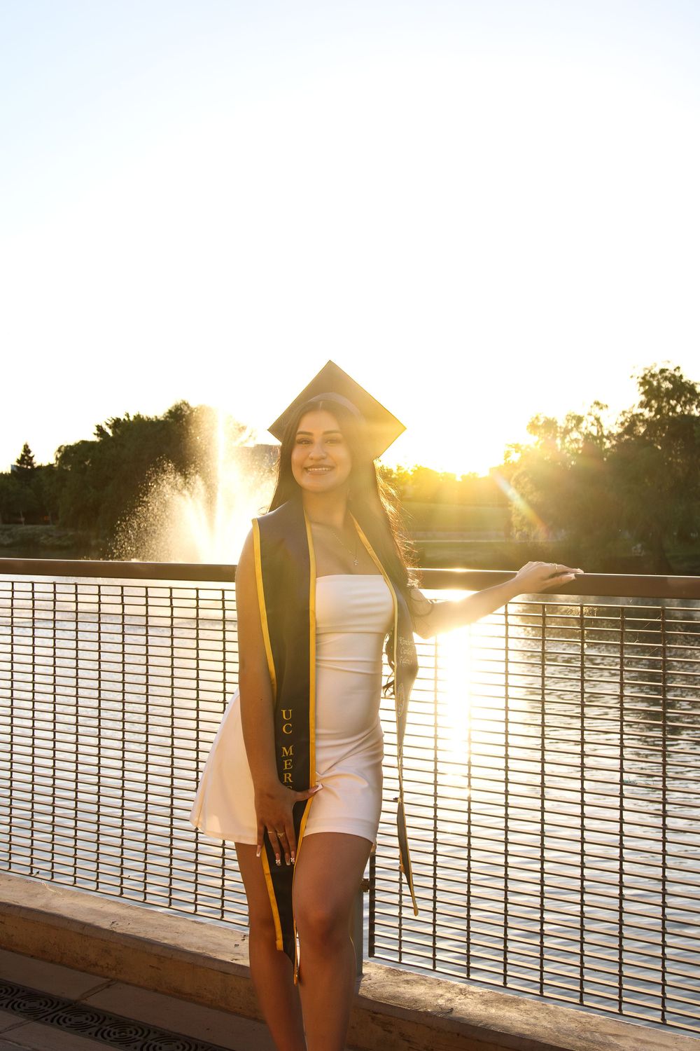 Graduate in cap and white dress posing by fountain at golden hour.