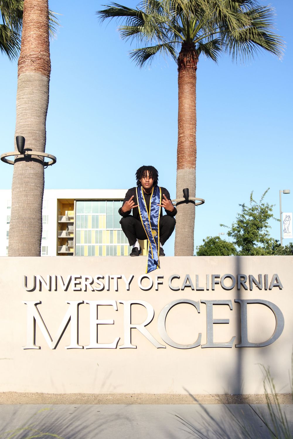 Graduate crouching on University of California Merced sign wearing blue and gold sash.