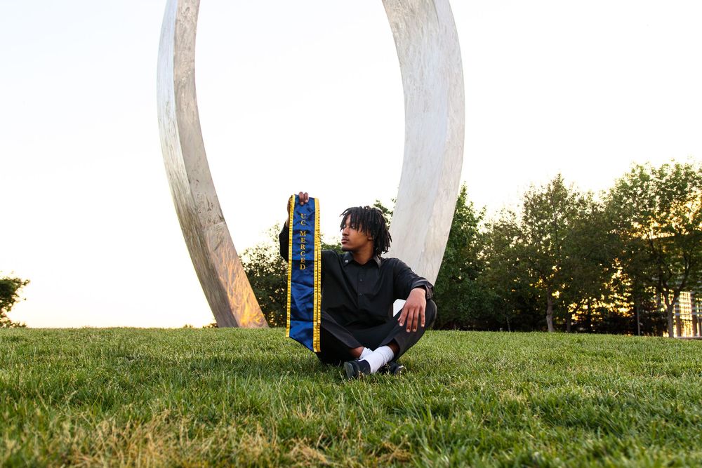 Young man sitting on grass beside a large circular metal sculpture outdoors.