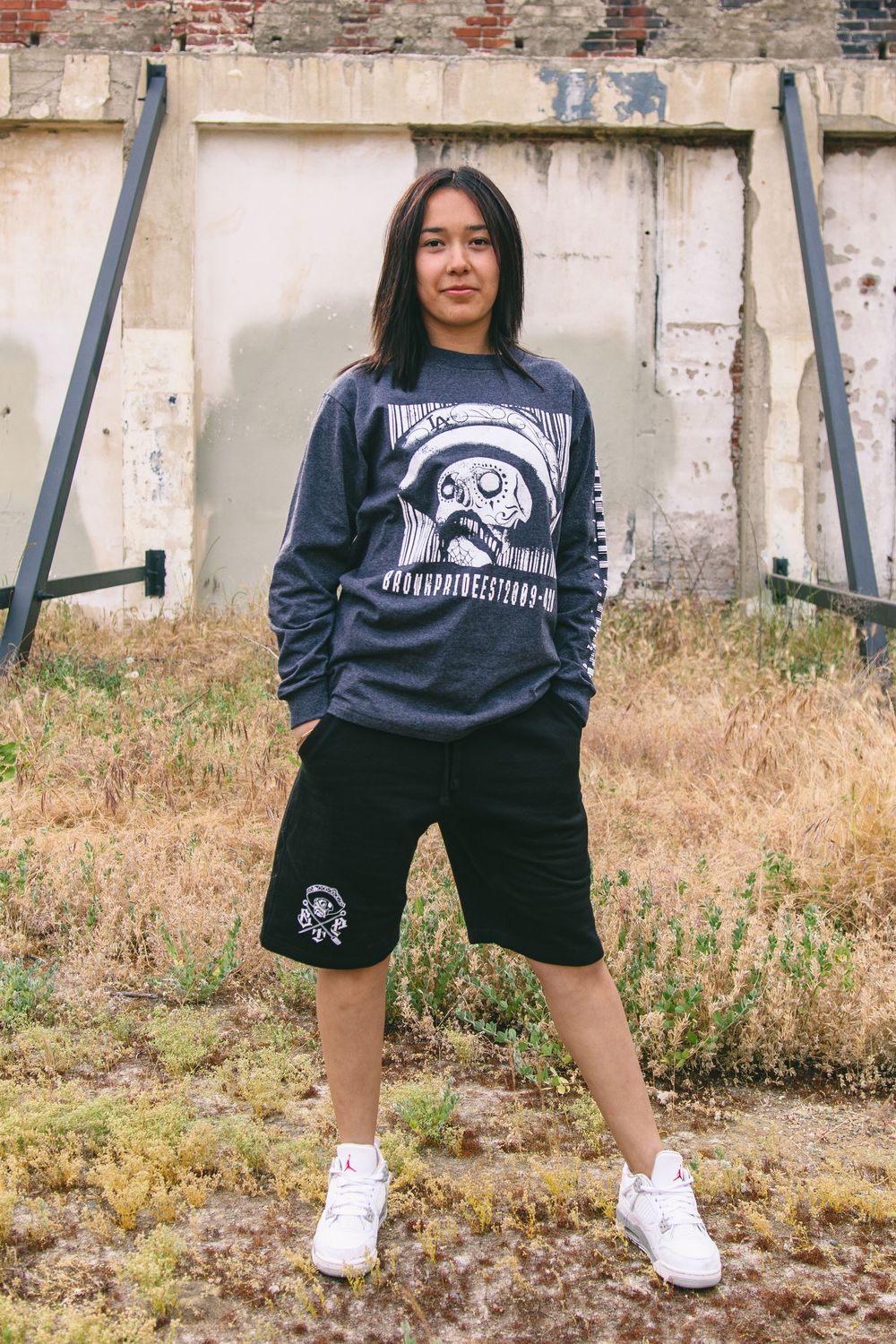 Teen in graphic sweatshirt and black shorts posing outdoors near worn concrete wall.