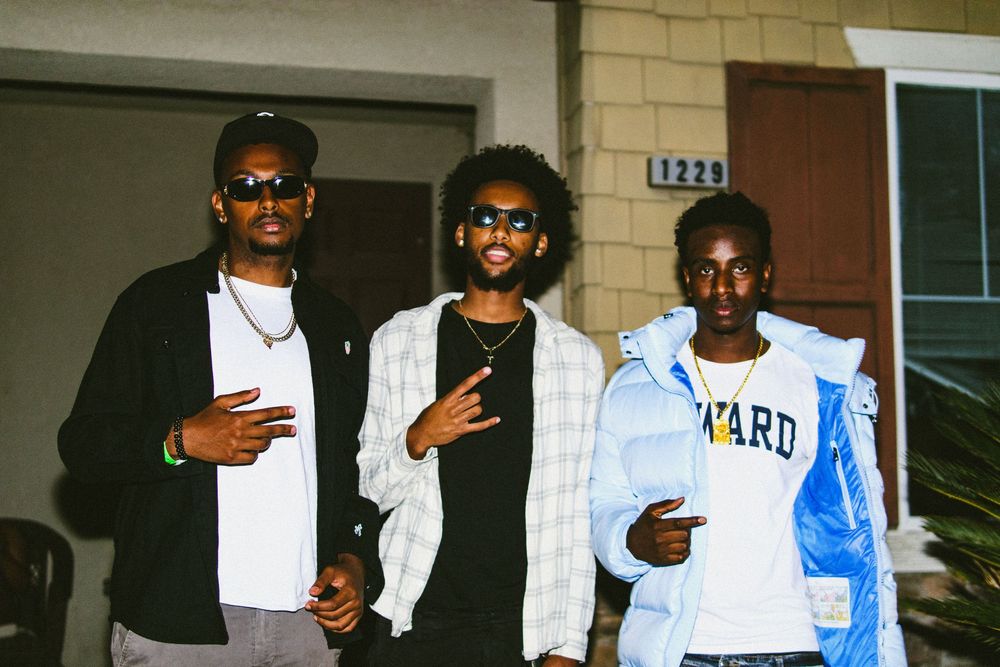 Three young men posing outside a house at dusk in casual streetwear.